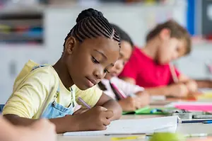 A girl in a classroom is sitting at a desk writing in a notebook with a pen