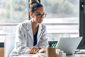 A woman wearing a white coat is working on a laptop inside a room with a glass window and trees outside