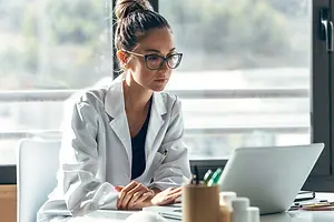 A woman wearing a white coat is working on a laptop inside a room with a glass window and trees outside