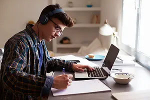A man is sitting on a chair and is looking at the laptop screen while writing on a notepad. He is wearing glasses and headphones. There are some books, a bowl, a plate, and a lamp on the desk. Behind him is a shelf with several items on it. There is a glass window on the right side. There is a reflection of the man in the window.