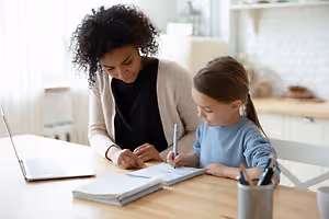 A woman and a girl are sitting at a desk in a kitchen, with the woman helping the girl write in a notebook