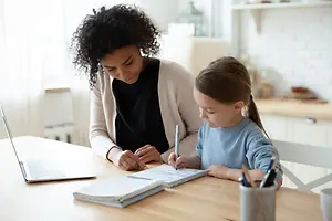 A woman and a girl are sitting at a desk in a kitchen, with the woman helping the girl write in a notebook