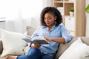 A woman is reading a book while sitting on a couch with white pillows.