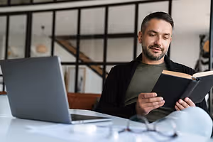 A man is reading a book while sitting on a chair in front of a table with a laptop.