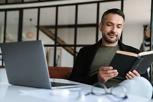 A man is reading a book while sitting on a chair in front of a table with a laptop.