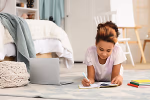 A girl is lying on the floor in a bedroom, holding a pen and writing in a book.