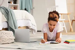 A girl is lying on the floor in a bedroom, holding a pen and writing in a book.