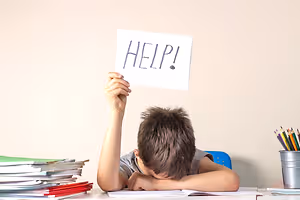 A young boy holding a sign that says HELP while sitting at a desk in a classroom