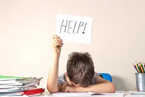 A young boy holding a sign that says HELP while sitting at a desk in a classroom