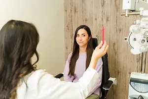 A woman sits in a chair holding a red pen while another woman examines her eyes with a device