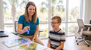 A woman and a boy are playing with toys in an office with glass windows and palm trees in the background