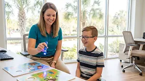 A woman and a boy are playing with toys in an office with glass windows and palm trees in the background