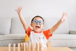 A child wearing glasses is raising his hands while playing a wooden game on a table.