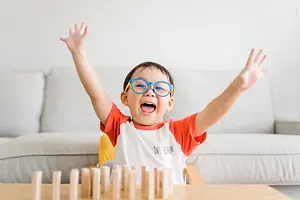 A child wearing glasses is raising his hands while playing a wooden game on a table.
