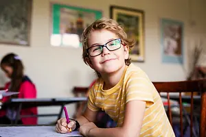 A smiling young boy with glasses is sitting on a chair in a classroom, holding a pencil, and seems to be drawing on a piece of paper.