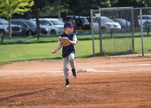 A young boy in a baseball uniform is holding a ball and standing on a baseball field. Behind him is a metal fence with a grassy area. On the other side of the fence are several parked cars, one of which is a white SUV. There are also trees and bushes in the area. The boy is wearing a baseball glove, glasses, and sneakers. The area is surrounded by grass. In the distance, there are some people standing near the cars.