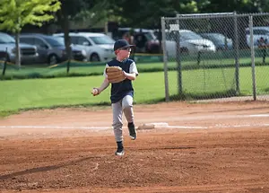 A young boy in a baseball uniform is holding a ball and standing on a baseball field. Behind him is a metal fence with a grassy area. On the other side of the fence are several parked cars, one of which is a white SUV. There are also trees and bushes in the area. The boy is wearing a baseball glove, glasses, and sneakers. The area is surrounded by grass. In the distance, there are some people standing near the cars.