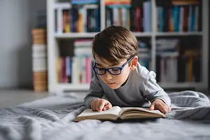 A young boy with glasses is reading a book on a bed with a white blanket and a shelf with many books in the background