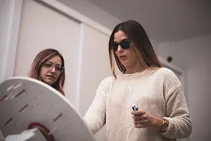 Two women with glasses and sunglasses in a lab, one holding a pipette and the other looking at a circular object on a wall