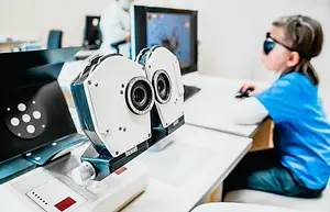 A girl in a blue shirt wearing glasses sits at a desk with a computer monitor and a machine resembling a camera or eyepiece in front of her.