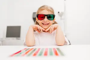 A young girl is smiling and wearing 3D glasses while sitting on a chair in front of a table with colorful objects on it.
