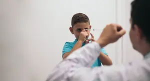 A doctor administers an eye drop to a young boy in a blue shirt in a room with a white wall.