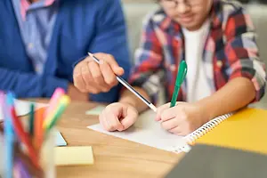 A child wearing glasses is writing on a paper with a pen while an adult is holding a pencil near the child.