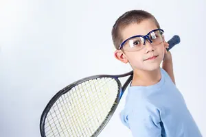 A boy wearing a blue shirt and glasses is holding a tennis racket and posing for a photograph