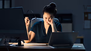 A woman sitting at a desk with her hands on her head while wearing glasses and a blue shirt.