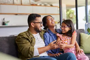 A family of three sitting on a couch, smiling and looking at each other, with one child wearing glasses