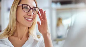 A smiling woman wearing glasses is holding her glasses up in front of her face