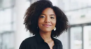 A woman with curly hair and a black top is smiling in front of a building with a glass wall.