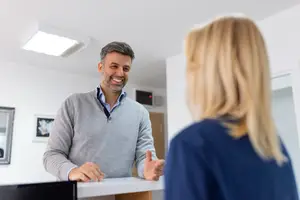 A smiling man in a gray sweater is standing in front of a desk and appears to be speaking with a woman in a blue shirt.