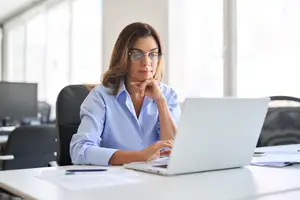 An adult woman is sitting on a chair in front of a desk with a laptop, wearing glasses and a blue shirt. She is looking at the laptop while smiling and touching her chin with her left hand. On the desk, there are some papers, a pen, and a monitor. Behind her, there is a window with a view of the outside. In the distance, there is a blurry view of another desk and chair.
