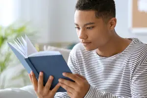 A young man wearing a striped shirt is sitting on a couch and reading a book
