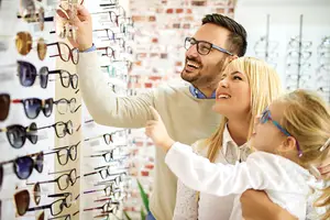 A family of three, including a father, mother, and daughter, are selecting glasses in a shop with a brick wall in the background.