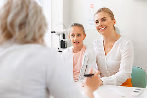 A woman and a young girl sitting in front of a table, probably in a clinic, and the woman is looking at the girl while smiling