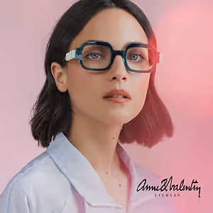A woman wearing Anne & Valentiny eyeglasses poses in front of a pink background