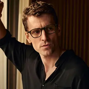 A man wearing a black polo shirt and glasses with a serious expression, standing in a room with wooden walls and looking at something in front of him.