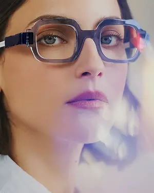 A woman with brown hair is wearing clear glasses with a silver frame, and she has makeup on, including eyeliner, lipstick, and a highlighter. She has a serious expression, and her face is slightly blurry, with the background behind her being a bit out of focus.