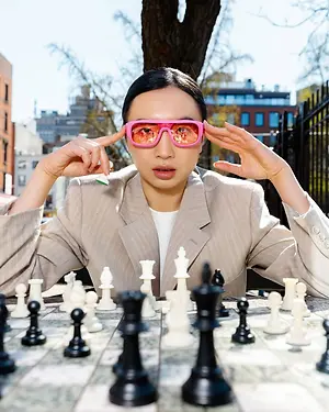 A woman wearing sunglasses is sitting on a bench and playing chess. She is looking at the chessboard and seems to be thinking deeply. Behind her is a blurry view of a fence, a tree, and a building with glass windows.
