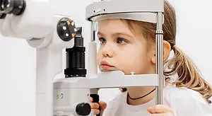 A young girl is looking into an ophthalmoscope, her hair tied in a ponytail, wearing a white shirt.