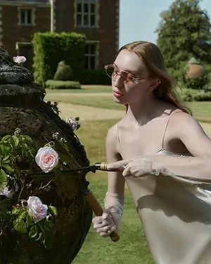 A woman in a white dress with glasses and gloves is trimming a rose plant growing on a stone vase in a garden with a mansion in the background.
