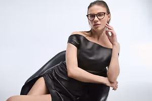A woman in a black dress and glasses is sitting on a black leather chair posing for a photo.