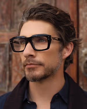 Man wearing black glasses with gold accents and a blue shirt standing in front of a wooden door