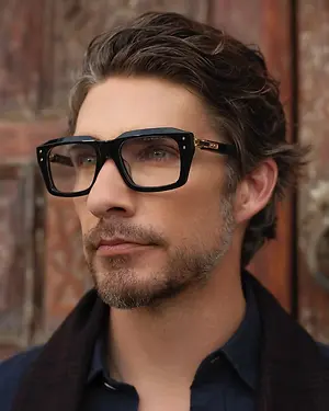 Man wearing black glasses with gold accents and a blue shirt standing in front of a wooden door