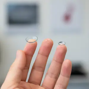 Three contact lenses on a finger in a room with a white wall and a blurry object in the background.