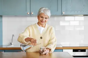 An older woman is pouring water into a glass in a kitchen.