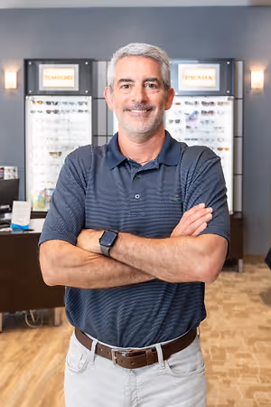 A man standing in front of a desk and a wall with two monitors. He is wearing a blue polo shirt and a belt. He is smiling and seems to be posing for a photo.