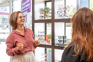 A woman wearing glasses is talking to another woman in a room with a glass wall and display of eyeglasses